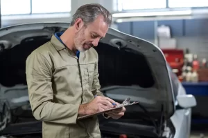 Imagem de homem branco de macacão verde em frente a carro fazendo checklist oficina mecânica.