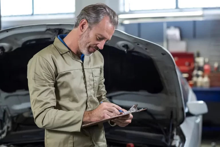 Imagem de homem branco de macacão verde em frente a carro fazendo checklist oficina mecânica.
