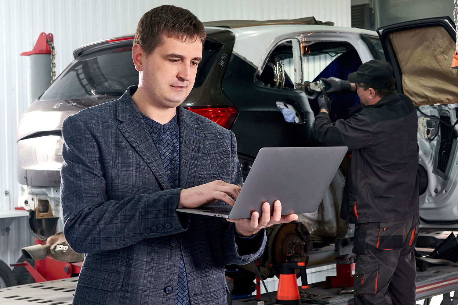 Homem de terno segurando um notebook e um mecânico trabalhando em um carro no fundo.