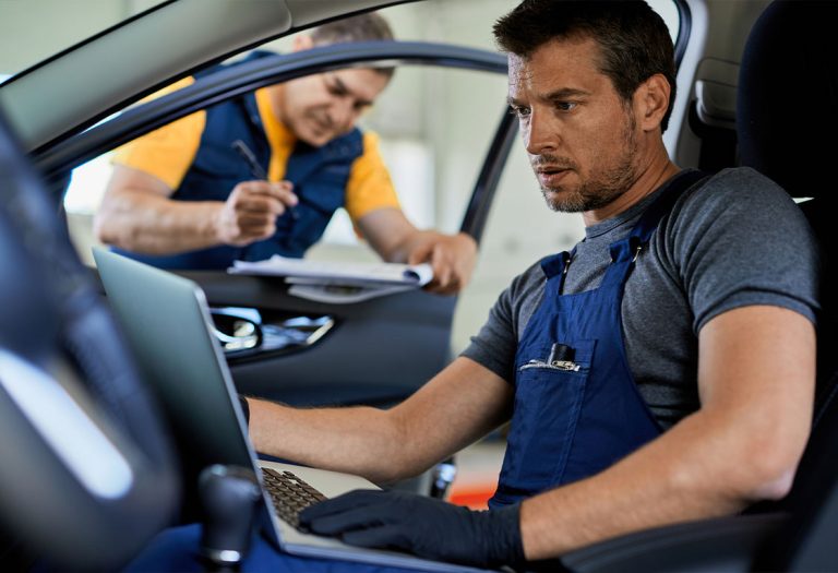 Homem dentro do carro com um notebook no colo, enquanto um homem está do lado de fora do carro escrevendo num papel.
