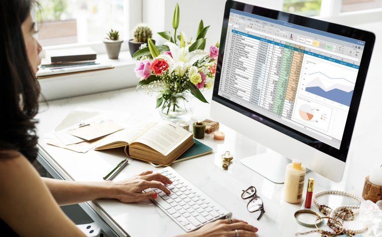 Mulher sentada em frente ao computador com uma planilha da ferramenta Excel aberta.