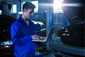 Mecânico segurando um notebook no pátio da oficina com dois carros atrás dele.